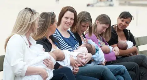 A group of 6 women breastfeeding their babies in a line on a park bench next to a river.